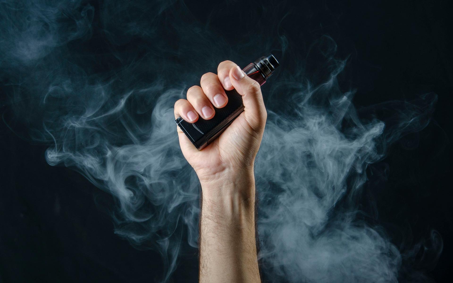 hand holding vape e-cigarette or electronic cigarette with white smoke over a black background.